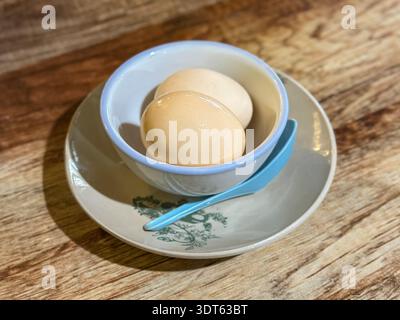 Two brown eggs placed inside a white bowl with blue rim on a floral saucer and spoon, rustic food arrangement on wooden table. Stock Photo