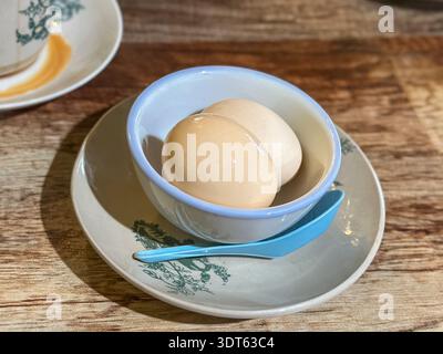 Two brown eggs placed inside a white bowl with blue rim on a floral saucer and spoon, rustic food arrangement on wooden table. Stock Photo