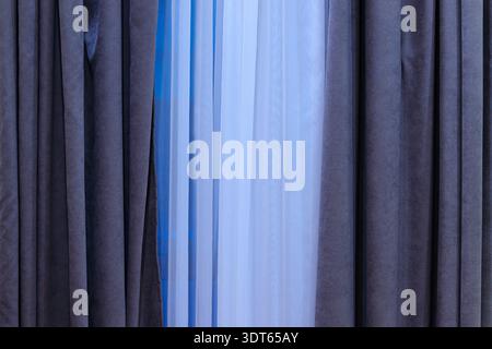 A close-up view of layered window coverings, with thick dark drapes framing delicate sheer textiles in white and blue hues Stock Photo