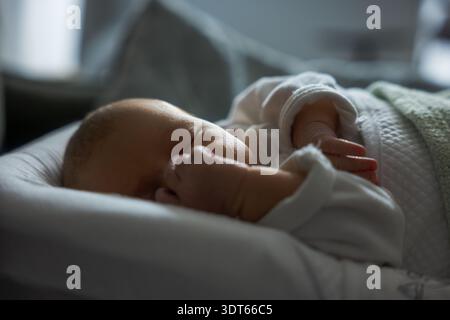 Newborn sleeps on a cushioned surface, white quilted onesie, tiny hands near face. Soft natural window light forms gentle shadows, pale green blanket Stock Photo