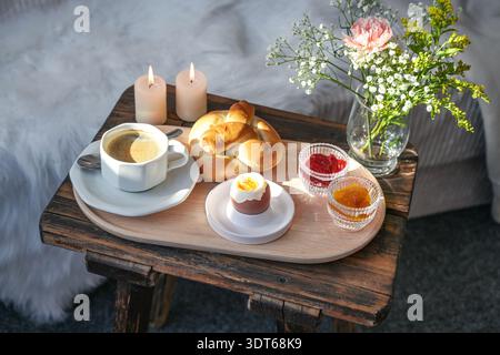 Breakfast prepared with love on an old rustic wooden bedside table in front of the bed, tray with coffee, jam, bun and egg, decorated with candles and Stock Photo