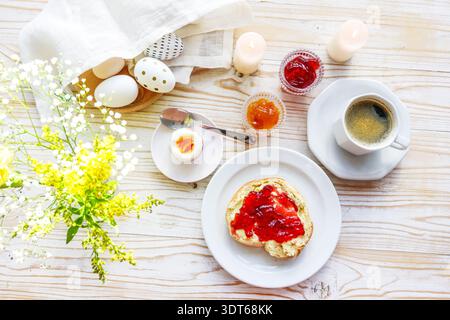 Easter breakfast, bun or sandwich with red jam, boiled egg, coffee, spring flowers and decorated eggs on a rustic light wooden table, top view from ab Stock Photo