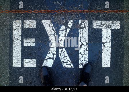 Painted white Exit sign on black concrete asphalt with two feet on top Stock Photo