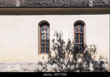 Two arched windows with wooden frames and decorative glass panels set in a weathered white wall beneath a wooden shingled roof. A tree shadow falls ac Stock Photo