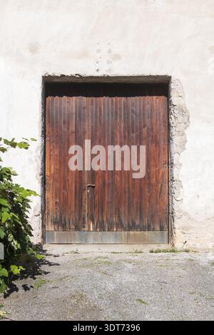 A weathered wooden garage door with vertical planks in warm brown tones is set against a white stucco wall. Green foliage appears on the left side of Stock Photo