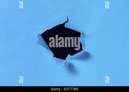 A hole in blue paper with torn edges and a black isolated background inside. Ripped blue paper, copy space Stock Photo