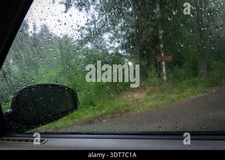 View through a car window covered with raindrops during summer rain, blurred forest road in the background Stock Photo