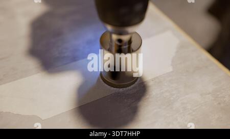 Worker drilling round hole into gray ceramic tile for plumbing installation, using diamond core bit on drill Stock Photo