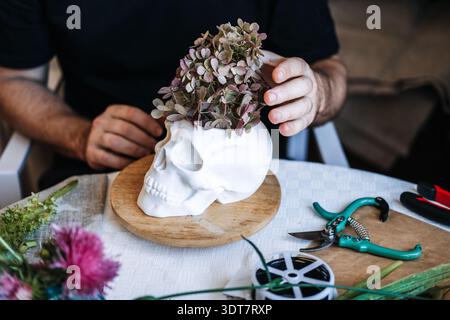 Hands place dried flowers into skull planter on table with floral tools. Moody home decor, gothic revival, maximalist interior design. Stock Photo