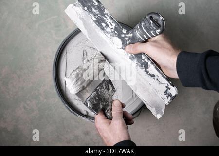 Top view of hands transferring wall filler from a putty knife onto a wide taping knife above a bucket. Concept of drywall finishing, wall repair and D Stock Photo