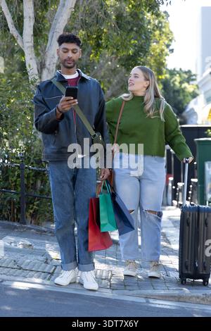 Couple walking on urban sidewalk, man holding smartphone, woman pulling suitcase with shopping bags Stock Photo