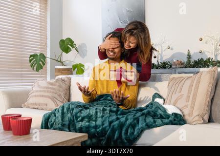 Indian and Asian couple on sofa in living room, woman giving wrapped gift to smiling man Stock Photo