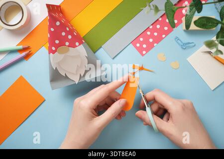 Hands cut carrots out of orange paper. Colorful paper, scissors, tape on blue background. DIY handmade art project for kids. Stock Photo