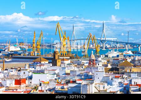 Cityscape showcases historic white rooftops of Cadiz, Spain, set against a modern industrial backdrop. Massive yellow shipyard cranes and the towering Stock Photo