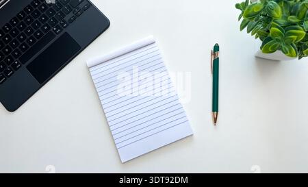 A blank notepad next to a pen, laptop, and houseplant on a white table. Concept for planning, ideas, notes, and workspace organization. Stock Photo