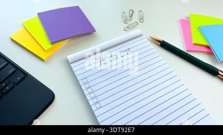 A notebook with the word "To Do List" written on it, a pen, sticky notes, and paper clips on a white table next to a laptop. Stock Photo