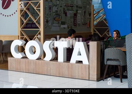 Large white 3D Costa Coffee brand signage in front of an indoor cafe seating area with customers relaxing in Abu Dhabi, UAE. Stock Photo