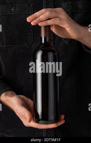 Dark glass wine bottle held by two hands against a corduroy-textured background. The bottle is cylindrical and lacks a label, featuring a smooth, refl Stock Photo