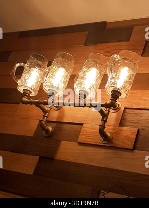 Warm vintage industrial wall light featuring four glass beer mug shades on metal pipe fixtures mounted to textured wooden paneling, casting cozy golde Stock Photo
