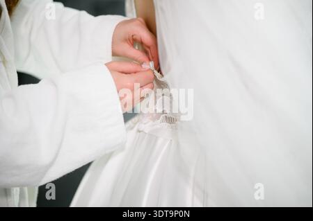 Bride getting ready. Tailor tries on wedding dress for woman, adjusting size to suit her. Bridesmaid carefully fastens delicate wedding dress in antic Stock Photo