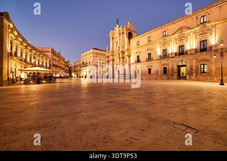 People enjoy dining outside in the square next to the Cathedral in Syracuse, Sicily. Lights illuminate the buildings as evening falls. Stock Photo
