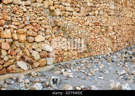 High-resolution shot of an ancient rustic stone wall made of natural rock standing on a sandy surface with scattered pebbles and organic debris. Stock Photo