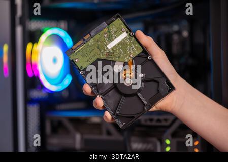 Hand holding opened hard disk drive in front of gaming computer with RGB lighting, internal storage component for data recovery and backup Stock Photo
