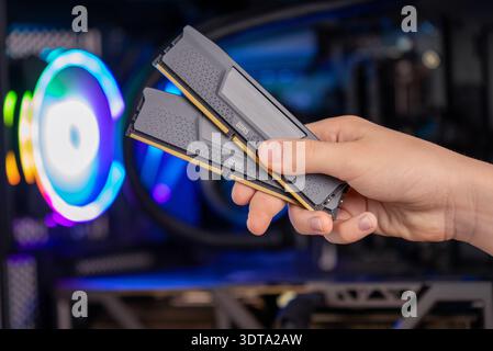 Hand holding two DDR5 RAM memory modules in front of gaming computer with RGB lighting, high performance system upgrade Stock Photo