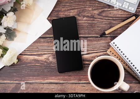 High angle view of smartphone on top of wooden desk with flower bouquet, computer keyboard, coffee and notepad Stock Photo