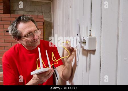 Connection of wifi router with repeater using LAN cable mature man adjusts equipment for internet distribution in rural house. Stock Photo