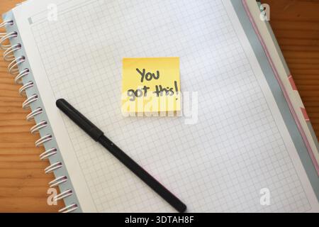 High resolution top view image of a yellow sticky note with the handwritten phrase “You Got This” placed on a grid notebook with a black pencil Stock Photo