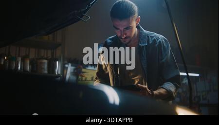 Young Auto Mechanic in Casual Wear Works in Fully Equipped Garage, Carefully Reviewing Information on Tablet. Man Uses Software Based AI For Diagnostics. Technical Inspection, Troubleshooting Process. Stock Photo