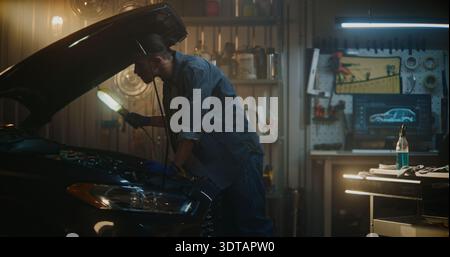 Man Stands in Well Equipped Garage Near Car With Hood Open, Conducting Technical Inspection Under Bright Service Lamp. Guy Examines Engine Components, Ensuring Proper Maintenance, Vehicle Performance. Stock Photo