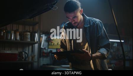 Young Man in Modern Workshop Focuses Intently on Tablet While Standing Next to Car With Open Hood. Male Mechanic Analyzing Data Before Proceeding With Repair. Concept of Professional Diagnostics. Stock Photo