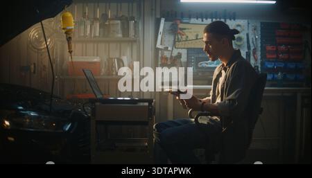 In Professional Garage, Male Technician Sits in Chair, Deeply Engaged in Analyzing Real Time Data on Tablet. Laptop on Mobile Workstation Also Displays Complex Diagnostic Graphs and Car Schematics. Stock Photo