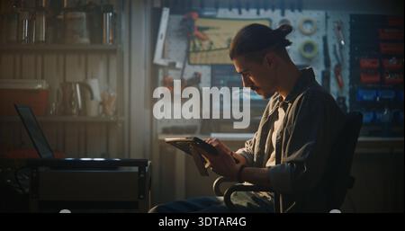 Male Automotive Specialist Seated in Workshop, Utilizing Dual Screen Setup With Laptop to Cross Reference Engine Data During Thorough Vehicle Inspection. Concept of Smart Diagnostic Solutions. Stock Photo