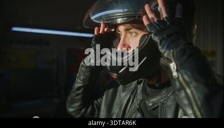 Close Up of Young Motorcyclists Face Prepares For Ride in Private Garage. Male Securing Chin Strap and Lowering Visor. Concept of State of Total Concentration Pilots Ready For Road. Slow Motion. Stock Photo