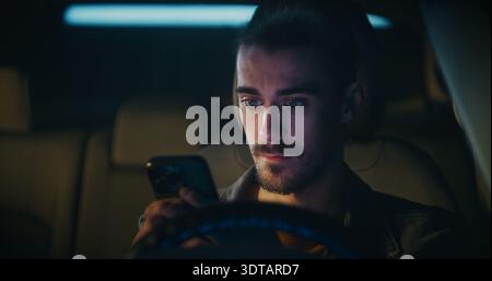 Young Man With Hair Tied in Bun Sits Behind Steering Wheel, Focused on Smartphone. With Determined, Male Types Long Message, Carefully Choosing Words. Emotional Involvement on Digital Communication. Stock Photo