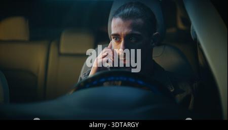 Guy Gets in Car Irritated While Continuing Unpleasant Phone Conversation on Smartphone. Sitting Behind Wheel, Speaks With Frustration. Work Stress, Emotional Tension, and Difficult Communication. Stock Photo