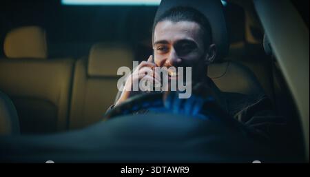 Young Man Sits Behind Wheel in Cheerful Mood, Talking on Smartphone and Laughing. Relaxed Guy Enjoys Lively Conversation. Concept of Positive Moment Modern Urban Lifestyle and Mobile Communication. Stock Photo