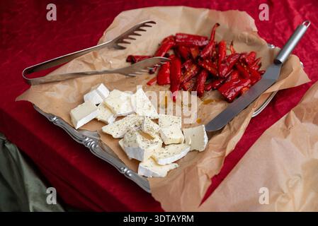 Close-up of a catering tray lined with brown parchment paper holding roasted red peppers and chunks of brie-style soft cheese sprinkled with herbs. Me Stock Photo