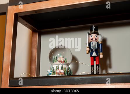 Festive holiday décor on a wooden shelf featuring a classic nutcracker figurine, a winter village snow globe and warm string lights against a neutral Stock Photo