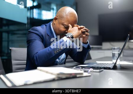 Black businessman sitting at desk with hands clasped near face, looking stressed Stock Photo