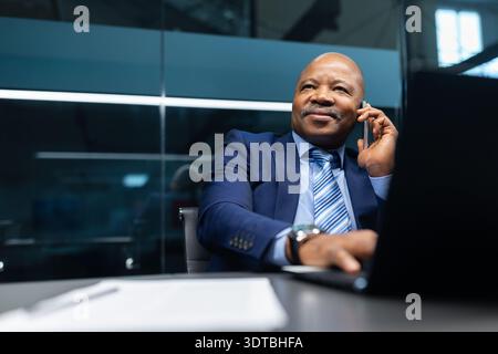 Confident mature black businessman talking on smartphone at desk in modern office Stock Photo