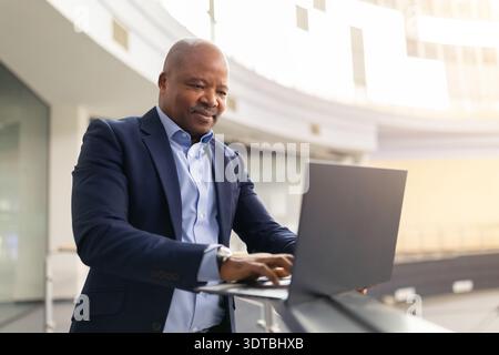 Focused mature black businessman working on laptop near office railing Stock Photo