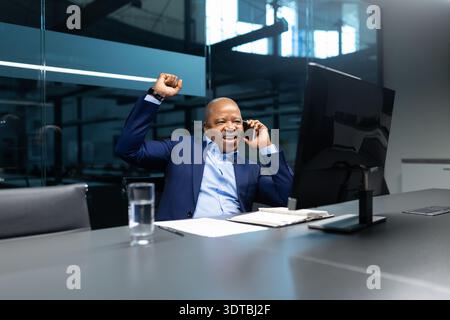 Excited black businessman celebrating success while talking on phone at office desk Stock Photo
