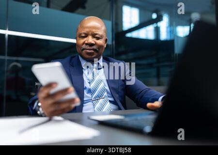 Mature black businessman sitting at desk using smartphone while working in office Stock Photo