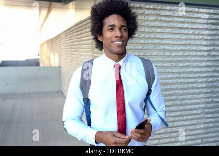 Young professional confidently standing outside with backpack and phone in morning sunlight Stock Photo
