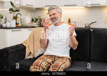 Middle-aged man at home records voice note on smartphone waves hello on speaker and shares news. Mature guy on sofa smiles talks sends chat reply then listens to playback nods and adds a short update. Stock Photo