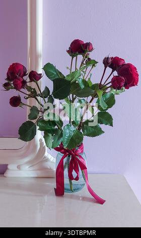 Dark Red Roses in a Glass Jar with Ribbon in front of a mirror. Romantic minimal still life with a cozy home mood on a soft pastel background, filled Stock Photo